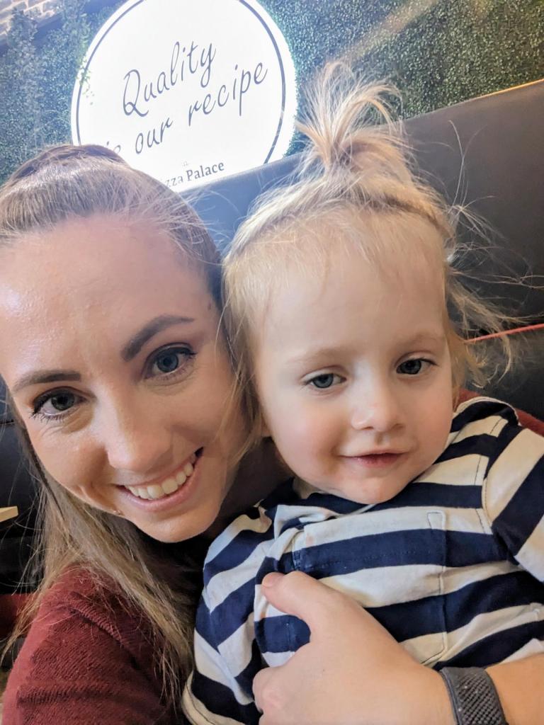 A smiling woman and a young child pose together for a selfie in front of a restaurant sign that reads 'Quality is our recipe' at Pizza Palace.